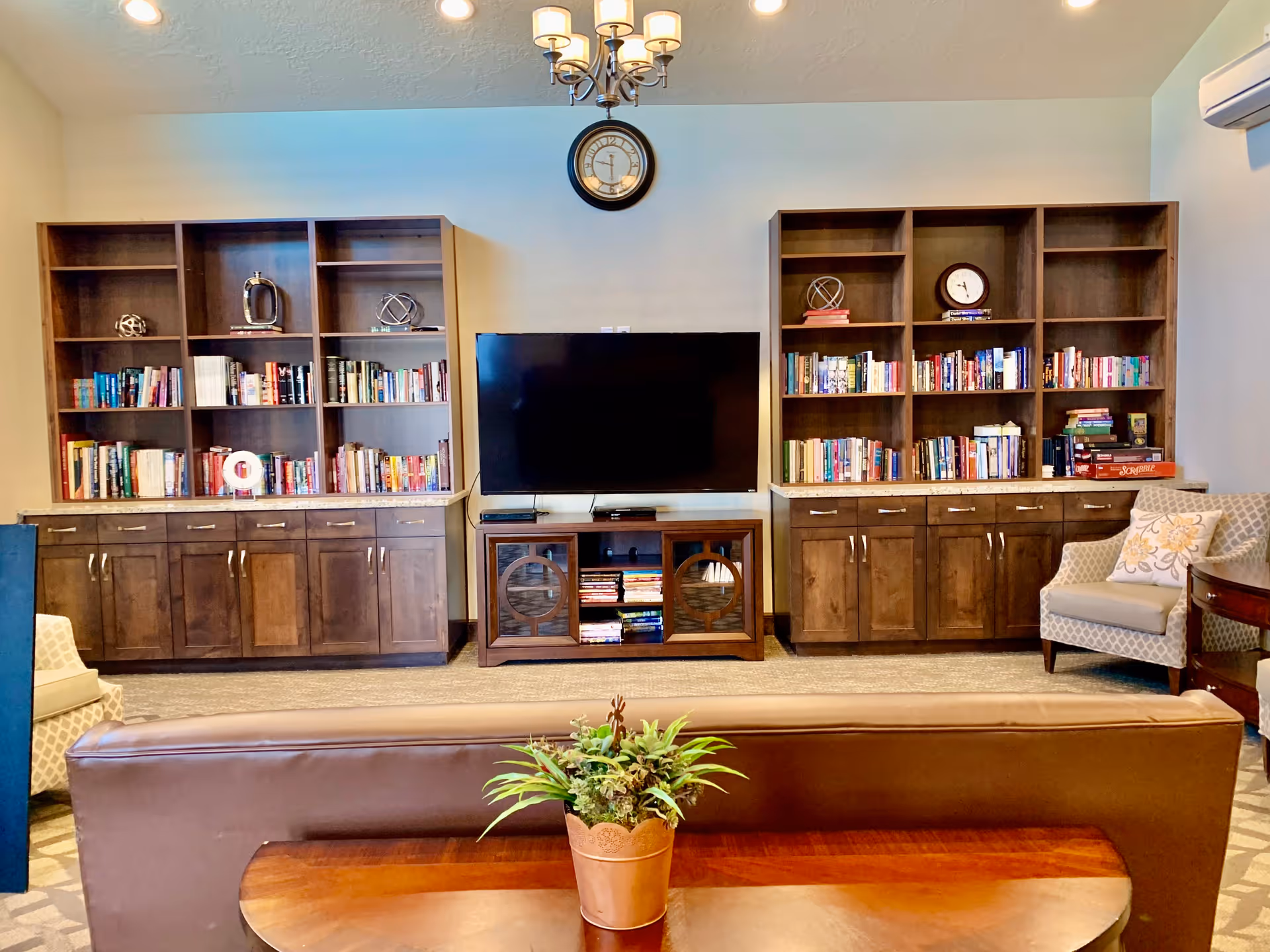 A cozy living room with two wooden bookshelves filled with books and decorative items flanking a flat-screen TV mounted on a wooden TV stand. Above the TV is a round wall clock and a chandelier with five lights. In the foreground, there is a brown leather sofa with a wooden table behind it holding a potted plant. To the right, there is a cushioned armchair with a patterned pillow and a small wooden side table.