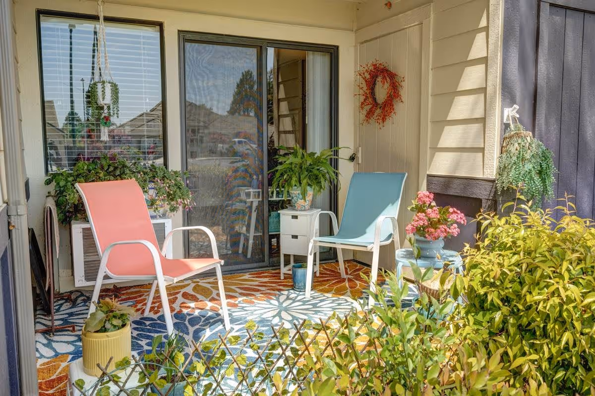 A cozy outdoor patio area with two colorful chairs, one pink and one blue, placed on a vibrant patterned rug. There are various potted plants and flowers around, including hanging plants and a wreath on the wall. The patio is adjacent to a sliding glass door and window with blinds.