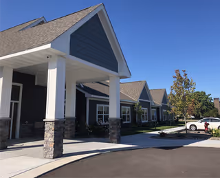Front entrance and exterior of a one-story senior living building with a covered portico supported by stone columns, landscaped grounds, and parked cars under a clear blue sky.