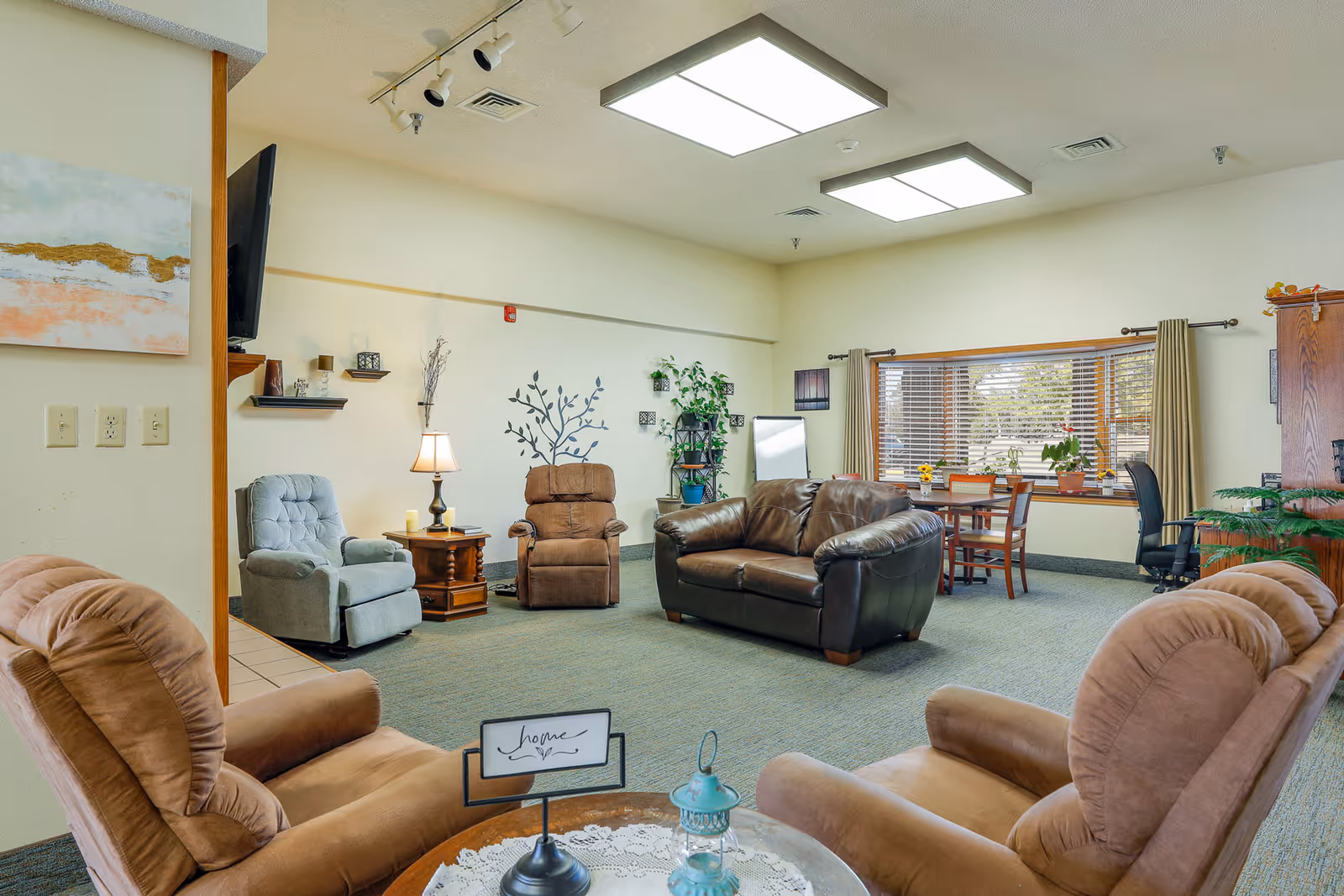 A cozy assisted living common area with multiple comfortable armchairs and a leather sofa arranged around a small table with a decorative sign reading 'home'. The room has a large window with blinds, a wooden dining table with chairs, plants, wall decorations, and soft lighting from ceiling fixtures and a table lamp.