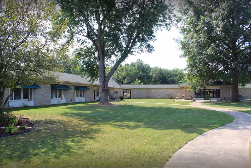 Single-story senior living building surrounding a grassy courtyard with large trees, a curved walkway, benches, and shaded windows.