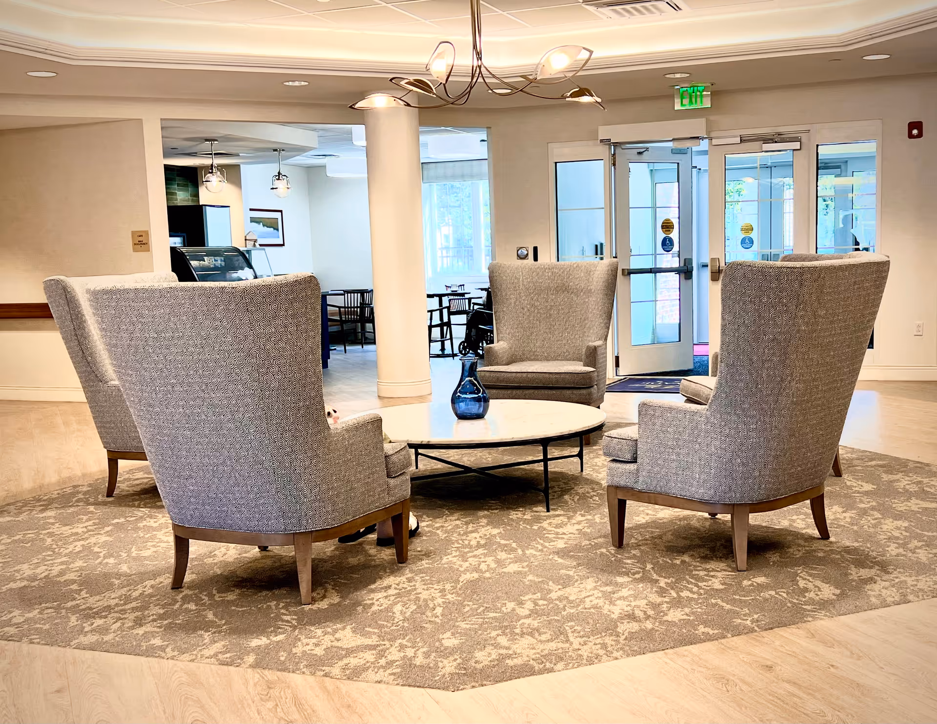 A cozy seating area in a senior living facility with four high-back upholstered armchairs arranged around a round marble coffee table with a blue vase on it. The area is carpeted with a patterned rug and is well-lit with ceiling lights. In the background, there is a doorway leading to another room with tables and chairs, and glass doors leading outside.