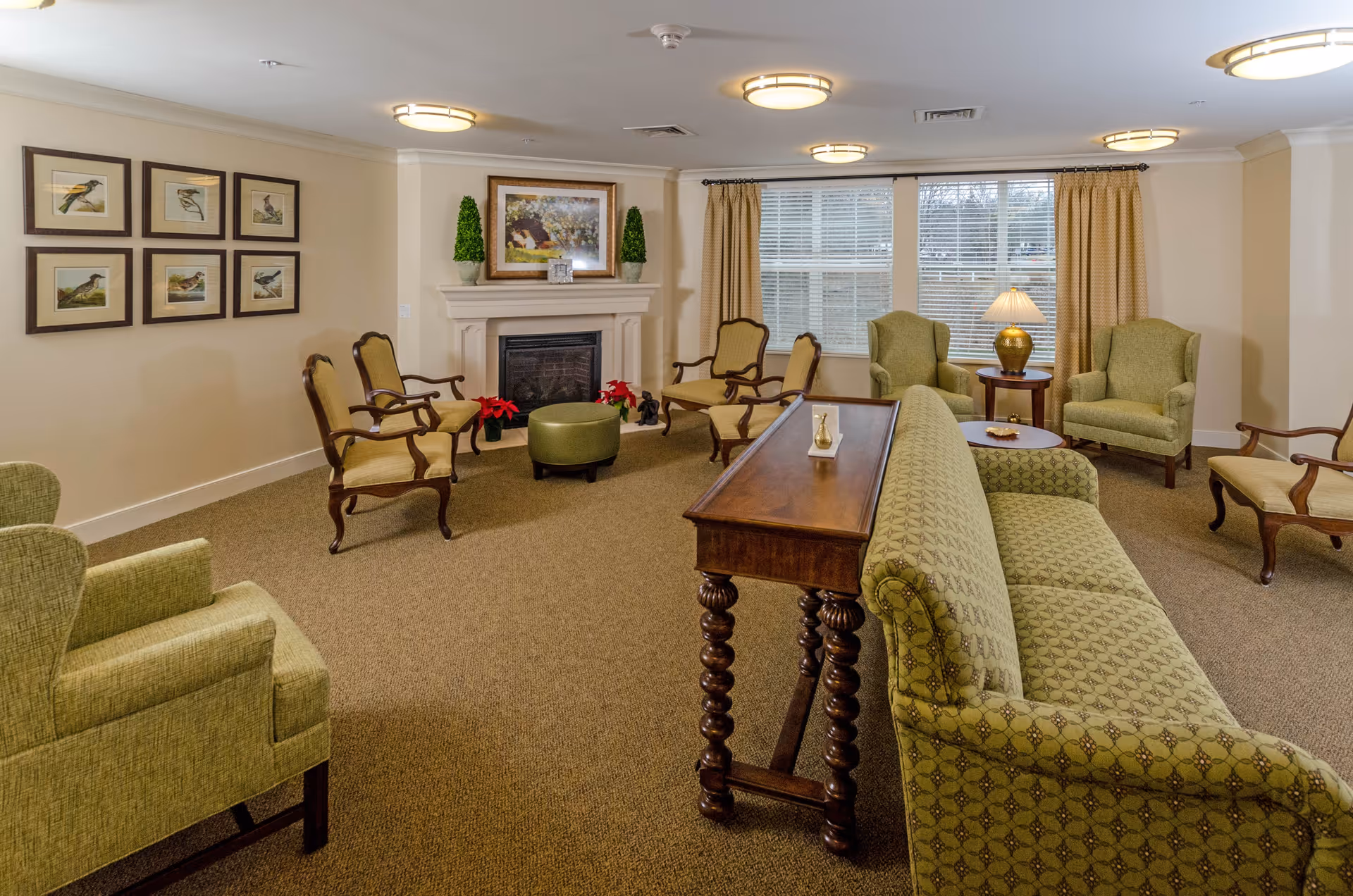 A cozy senior living facility common area with green upholstered armchairs and a matching sofa arranged around a wooden table. The room features a fireplace with decorative plants and a framed painting above it. There are six framed bird illustrations on one wall and large windows with beige curtains letting in natural light.