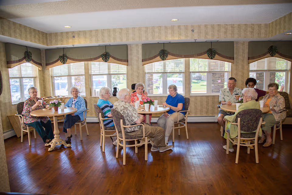 A group of seniors seated around several tables in a bright communal dining room.