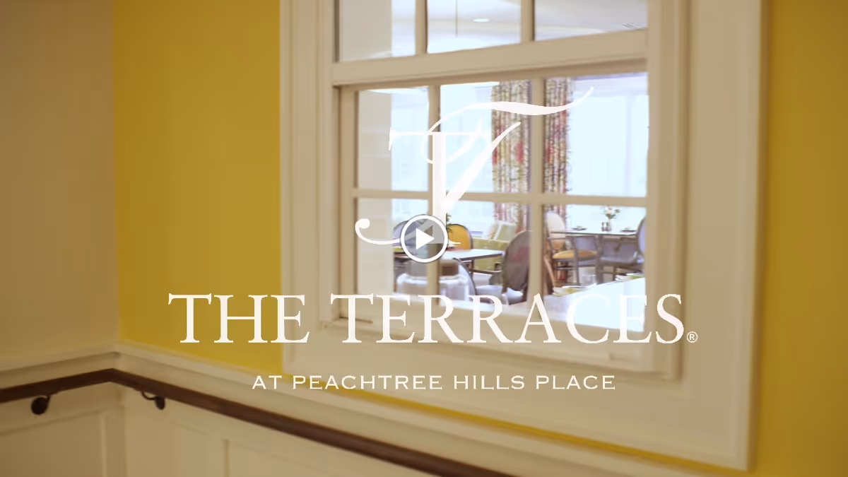 A yellow hallway interior window looking into a dining room with tables and chairs, overlaid by the text "THE TERRACES AT PEACHTREE HILLS PLACE".