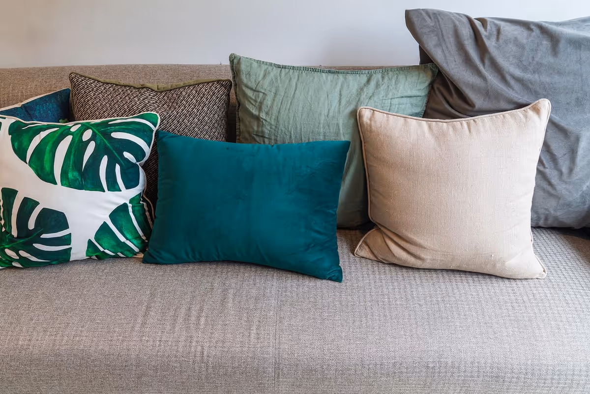 A beige fabric sofa with six decorative pillows in various colors and patterns, including green with leaf prints, solid teal, light green, beige, and gray.