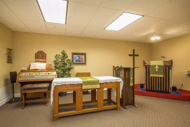 Interior of a small chapel or worship room with a wooden altar covered with a white cloth and green runner, a wooden lectern with a green cloth featuring a cross, a wooden pulpit on a red carpeted platform, a wooden organ with a bench, a potted plant, and framed pictures on beige walls.