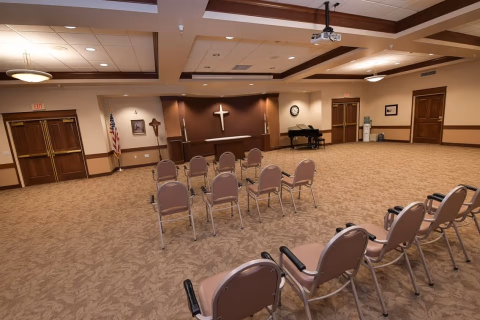 A spacious room with beige carpet and walls, arranged with chairs facing a small altar with a cross on the wall behind it. The room has wooden double doors, an American flag, a piano, a clock, and a water cooler. Ceiling lights and a projector are visible.