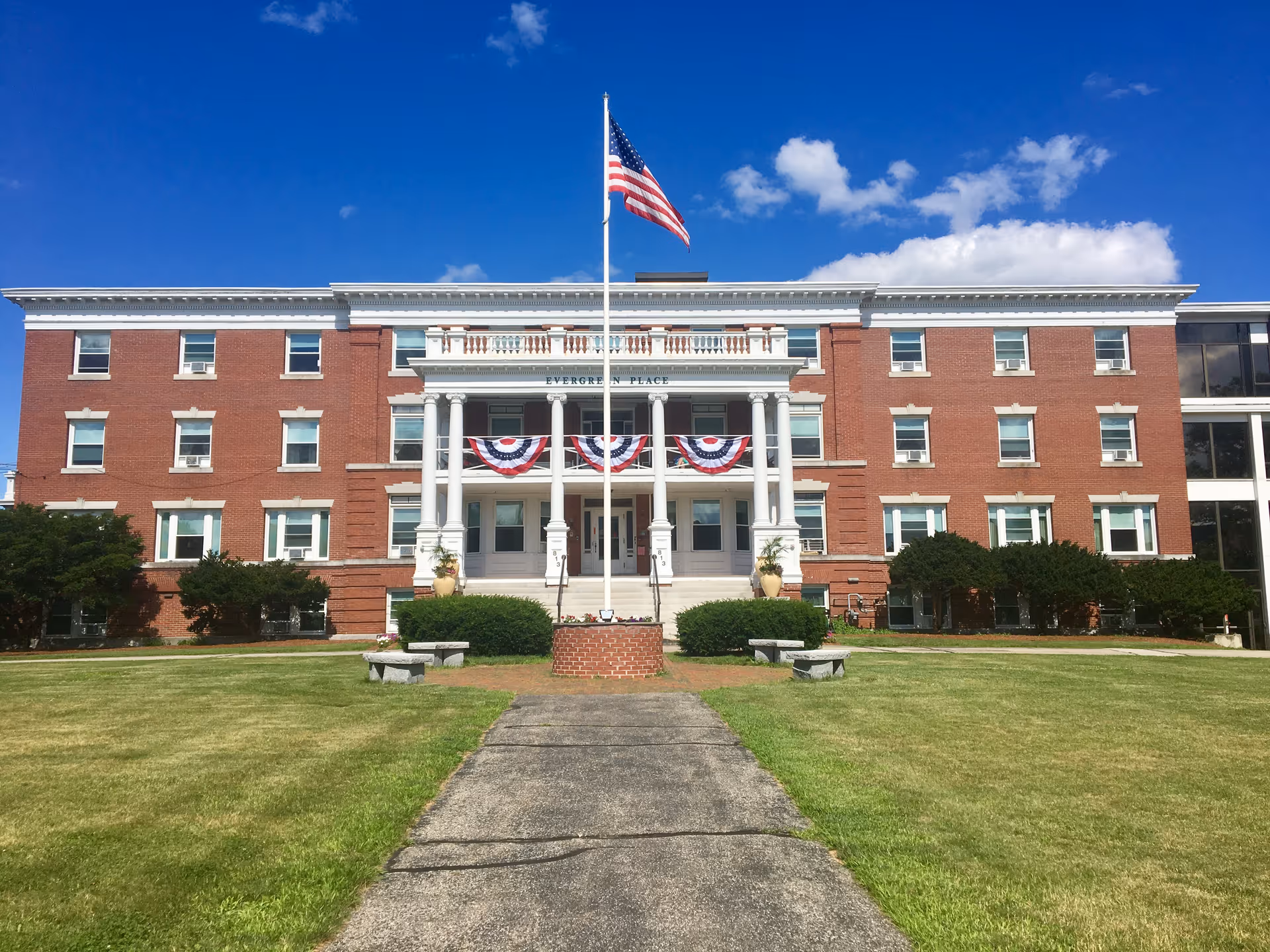 Front view of Evergreen Place, a three-story red-brick building with white columns, an American flag, and a paved walkway across a manicured lawn.