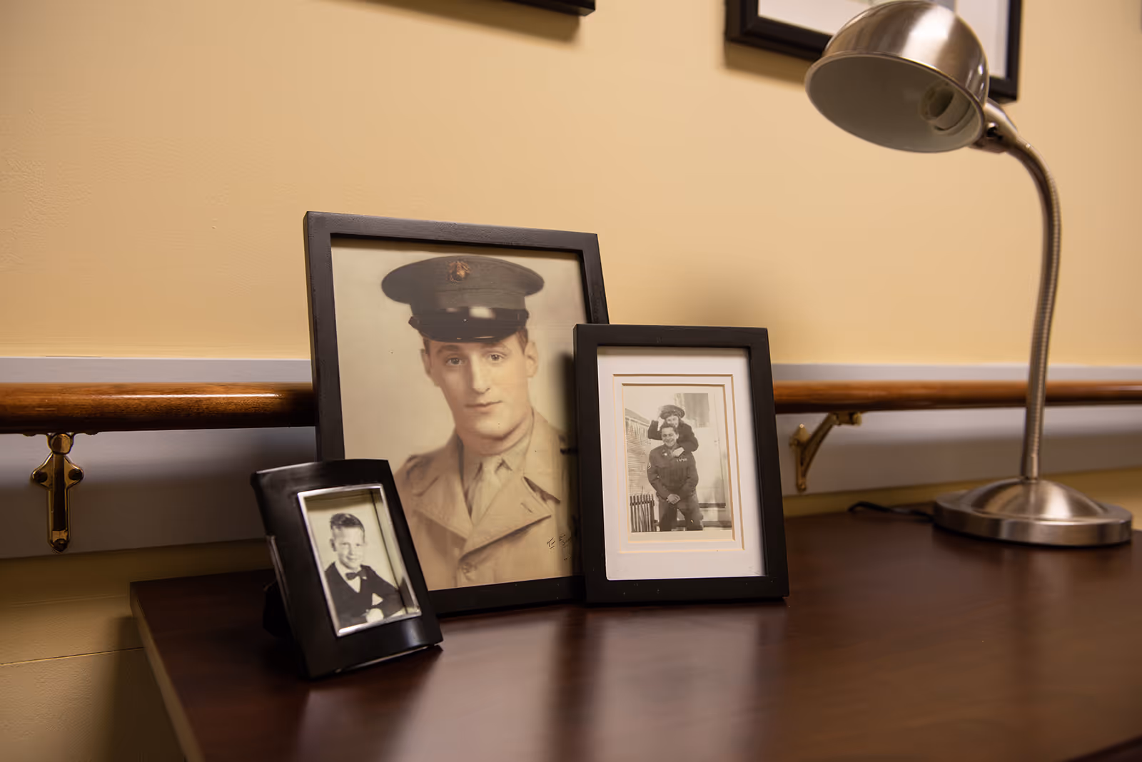 A wooden table with three framed black and white photographs, including a portrait of a man in military uniform, a smaller portrait of a young boy in a suit, and a photo of two people in military attire. A silver desk lamp is positioned on the right side of the table. The background features a beige wall with a wooden handrail.