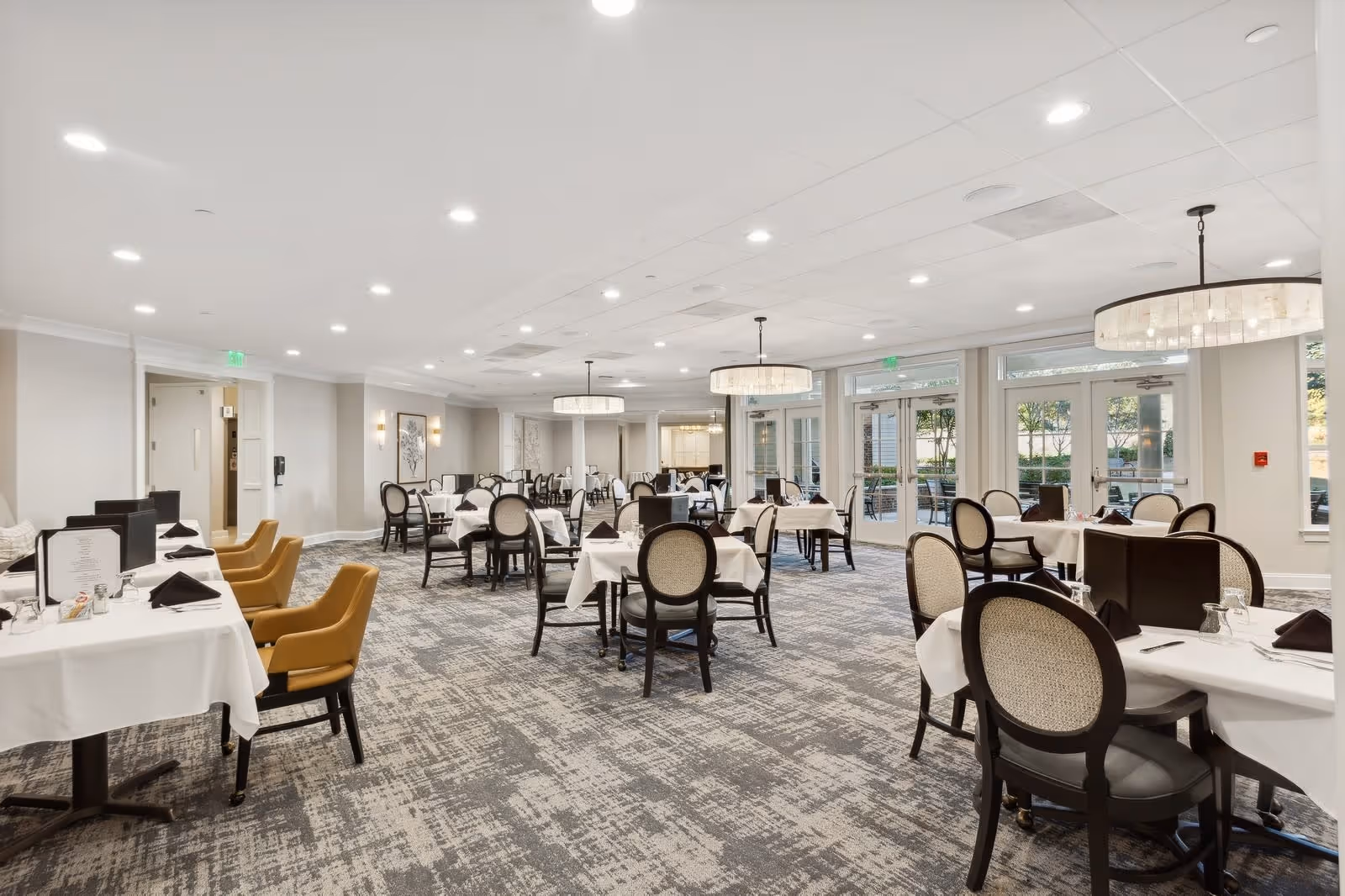 A spacious dining room with multiple tables covered in white tablecloths, each set with black napkins and glassware. The room features a patterned carpet, beige walls, large windows letting in natural light, and modern circular chandeliers hanging from the ceiling. There are a mix of black and tan chairs around the tables.