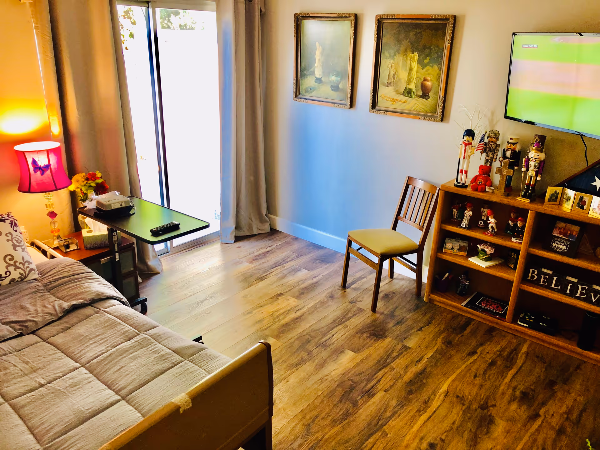 A cozy room with a hospital-style bed covered in a beige quilt, a bedside table with a purple lamp and flowers, a small adjustable table with a remote control and device on it, a wooden chair with a yellow cushion, two framed paintings on the wall, a wooden shelf filled with decorative items including nutcrackers and a folded American flag, and a wall-mounted TV showing a green screen.