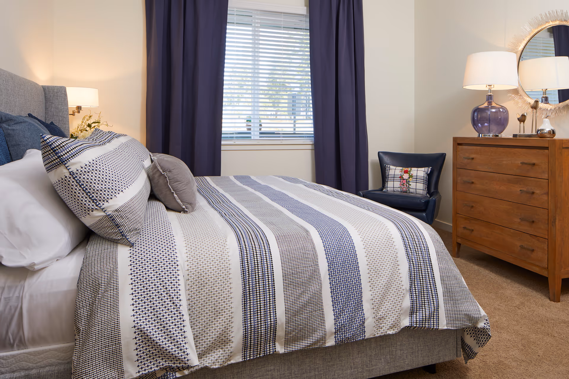 A neatly made bed with a striped blue, white, and gray comforter and matching pillows in a bedroom. The room features a window with closed blinds and dark blue curtains, a wooden dresser with a purple glass lamp and decorative items, a round mirror on the wall, and a dark blue chair with a decorative pillow.
