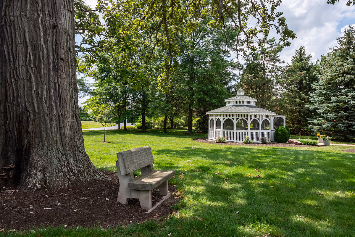 A peaceful outdoor garden area featuring a large tree with a stone bench at its base and a white gazebo surrounded by green grass and trees under a partly cloudy sky.