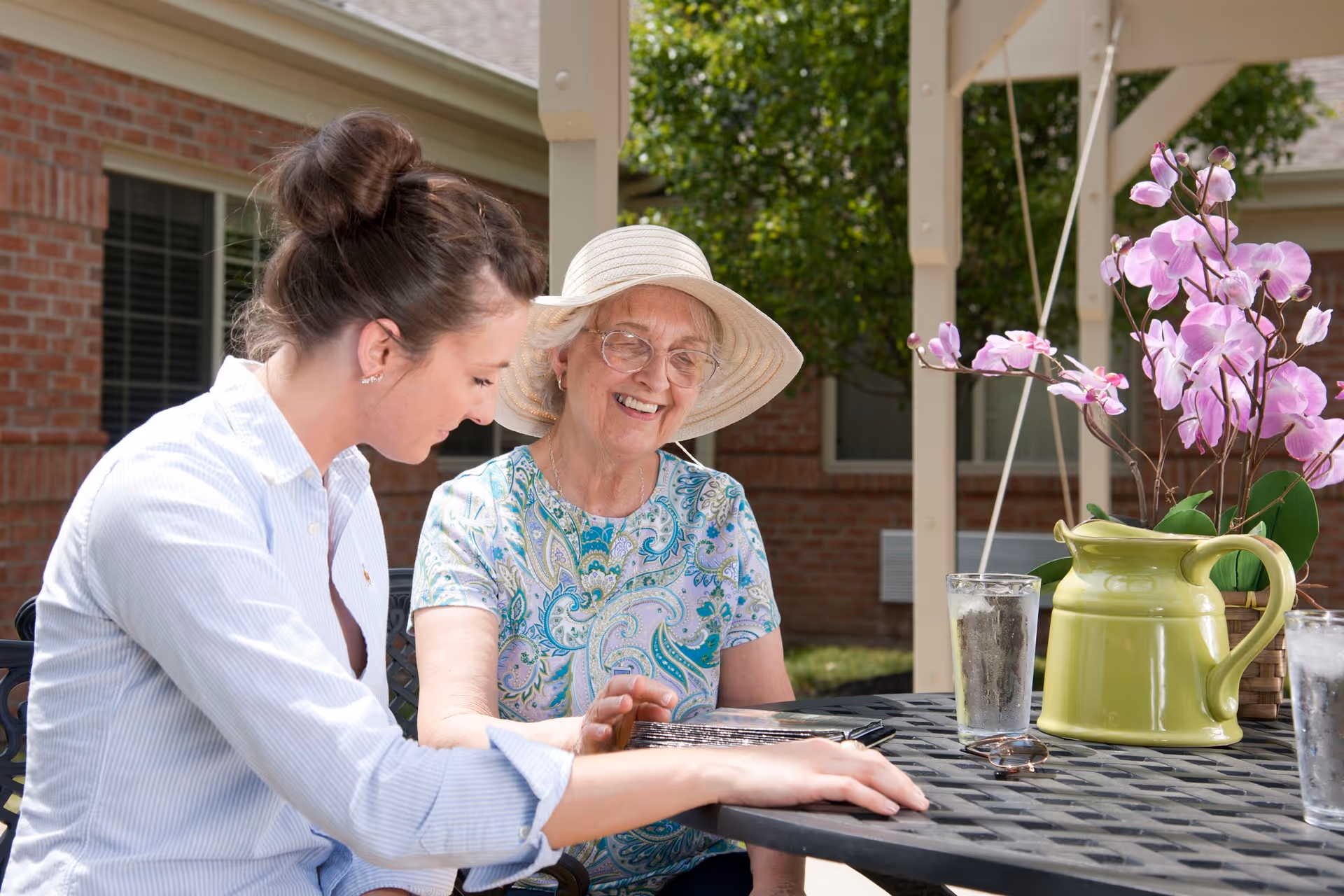 An elderly woman wearing a sunhat and glasses smiles while sitting at an outdoor table with a younger woman who is showing her something on a tablet. The table has a green pitcher with pink flowers and two glasses of water. They are seated under a covered patio with a brick building and greenery in the background.