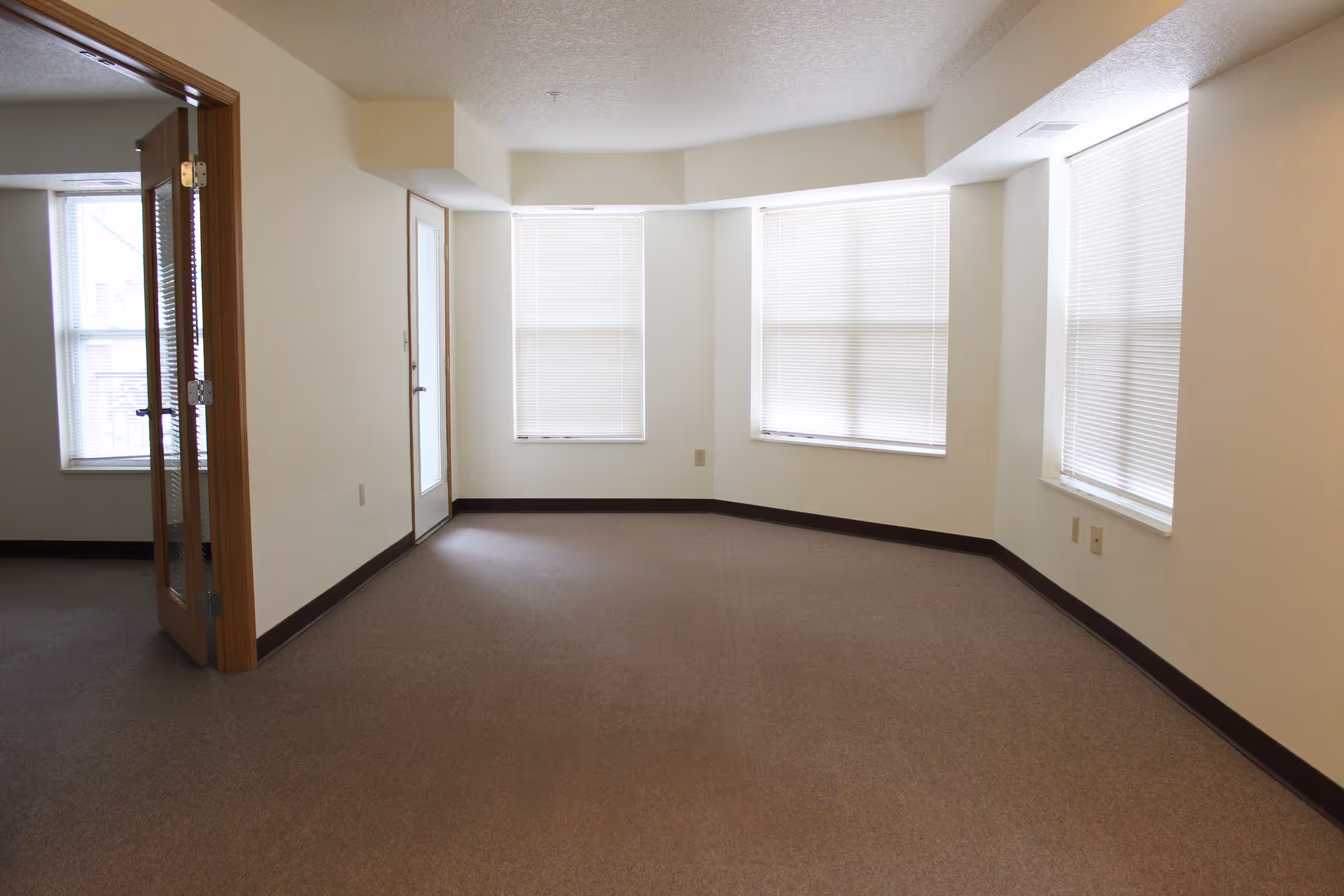Empty room with beige carpet and white walls featuring three windows with closed blinds. There is a wooden door with glass panels on the left side leading to another room with a window.