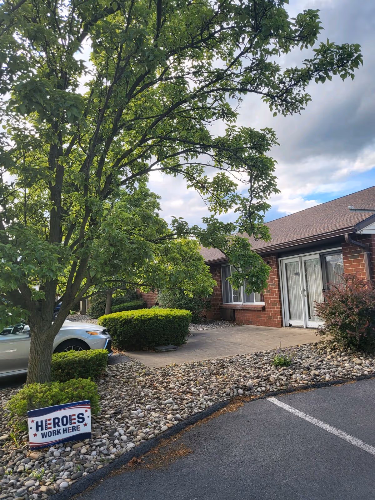 Exterior view of a brick building with a brown roof, surrounded by green trees and bushes. There is a paved walkway and a parking area with a silver car partially visible. A sign near the tree reads 'HEROES WORK HERE'. The sky is partly cloudy.