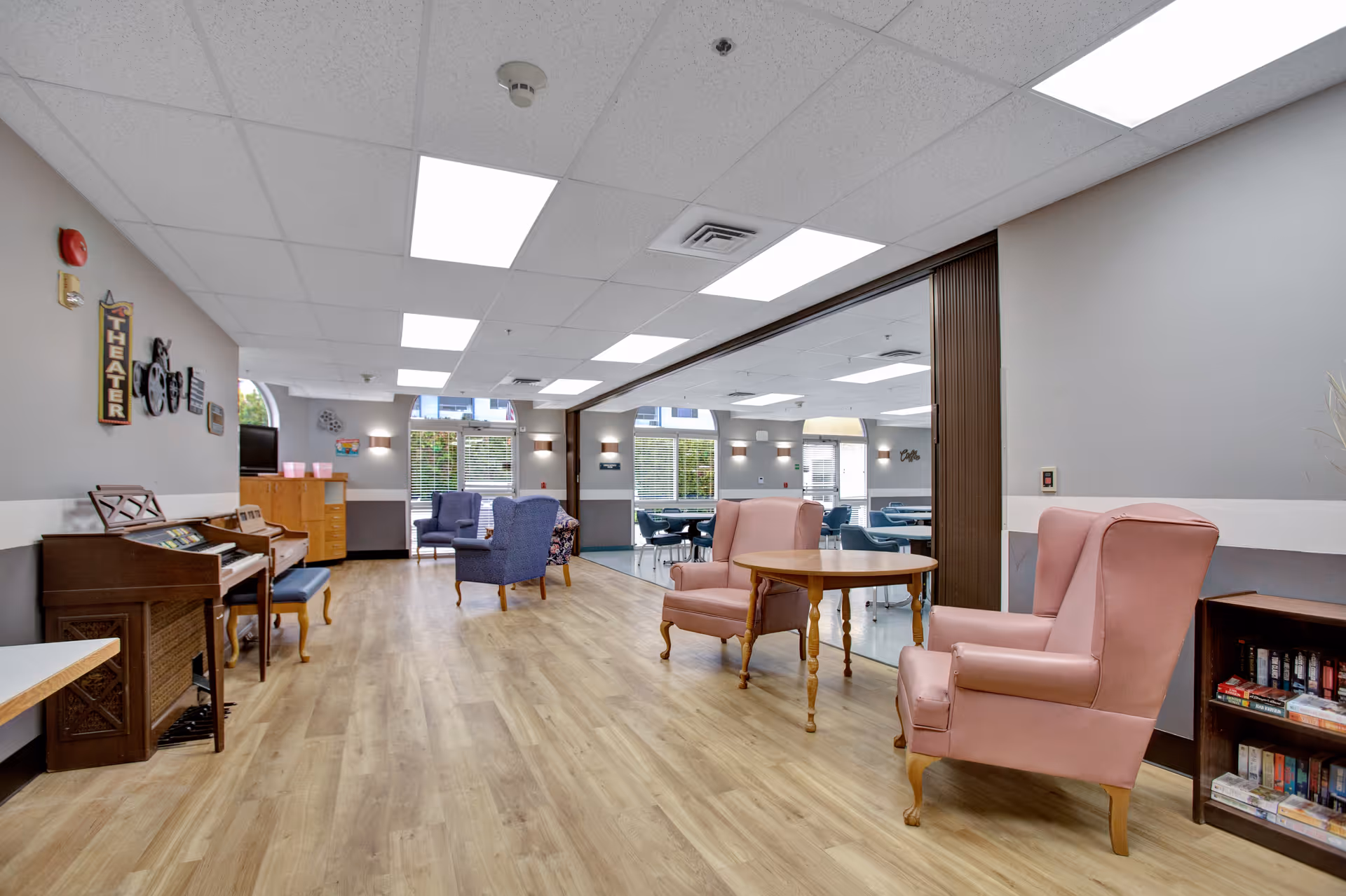 A spacious common area in a senior living facility with wooden flooring, pink armchairs around a wooden table, blue armchairs near windows, a piano, and a bookshelf filled with board games. The room is well-lit with ceiling lights and natural light from large windows.