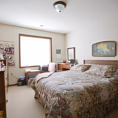A bedroom with a large bed covered in a patterned bedspread, a wooden headboard, a window with natural light, a chair with a pink blanket, a small desk with a mirror, and a framed picture on the wall.