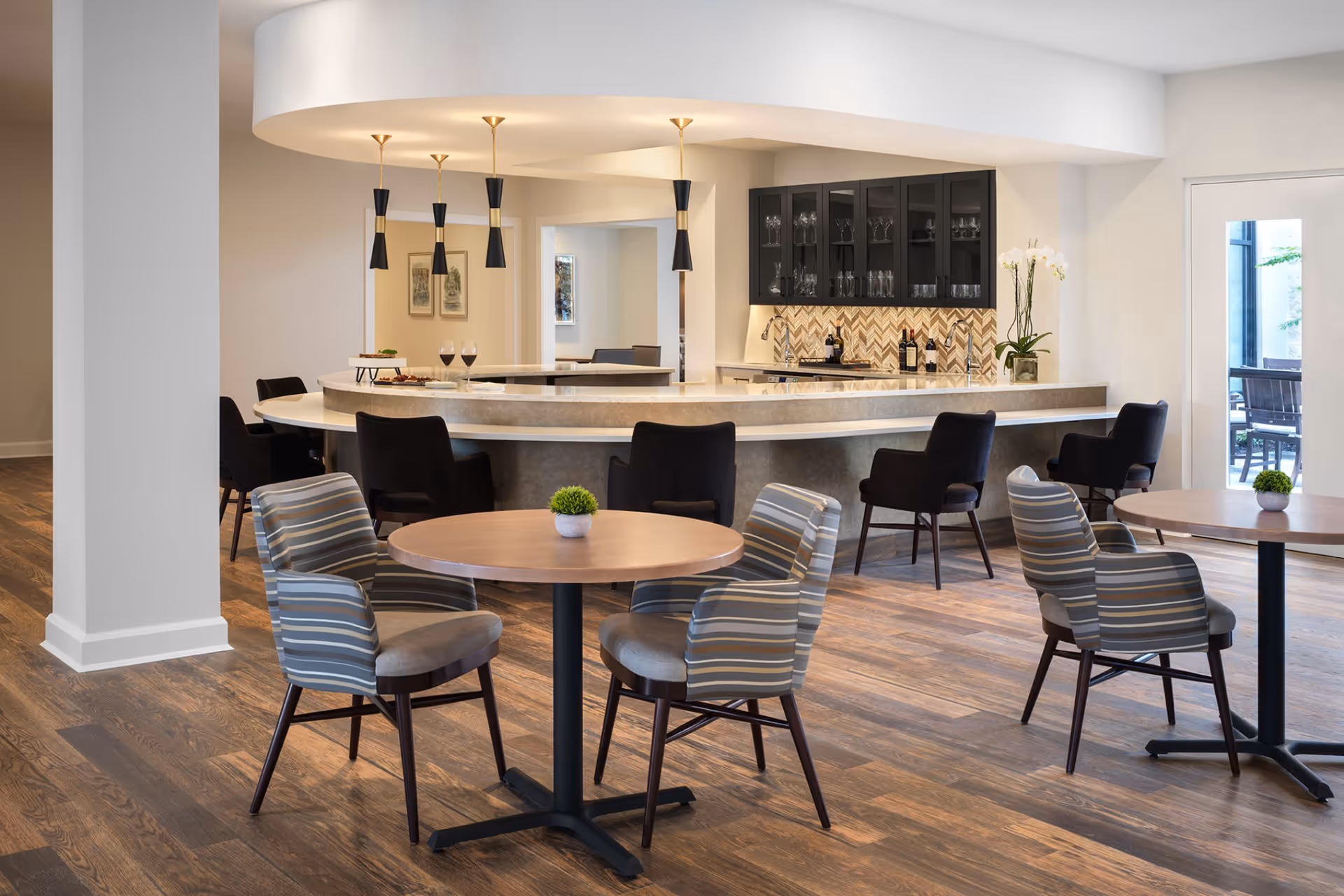 A modern dining area in a senior living facility featuring round wooden tables with striped cushioned chairs. In the background, there is a curved bar counter with black chairs, pendant lights hanging from the ceiling, and a cabinet with glassware. The room has wooden flooring and a door leading to an outdoor patio.