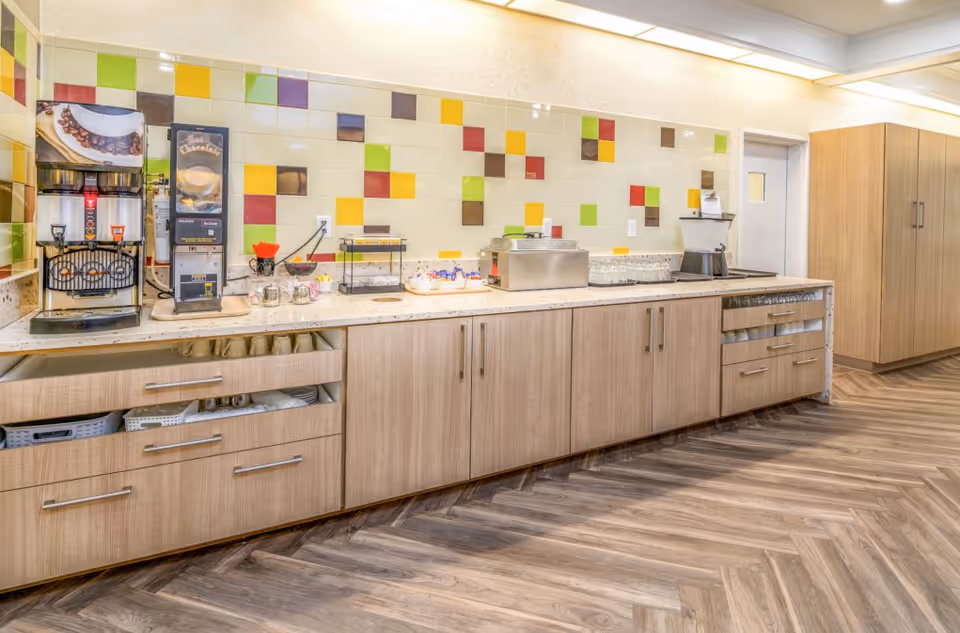 A bright kitchen area with a long countertop featuring coffee and hot chocolate dispensers, a toaster, and various condiments. The backsplash has colorful square tiles in green, yellow, red, and brown. Light wood cabinets and drawers are below the counter, and the floor has a wood-patterned herringbone design.