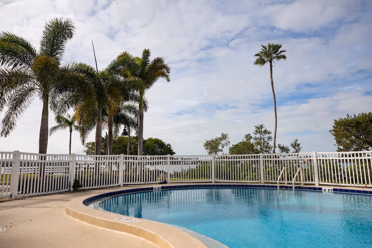 Outdoor swimming pool with clear blue water surrounded by a white fence, palm trees, and a partly cloudy sky in the background.