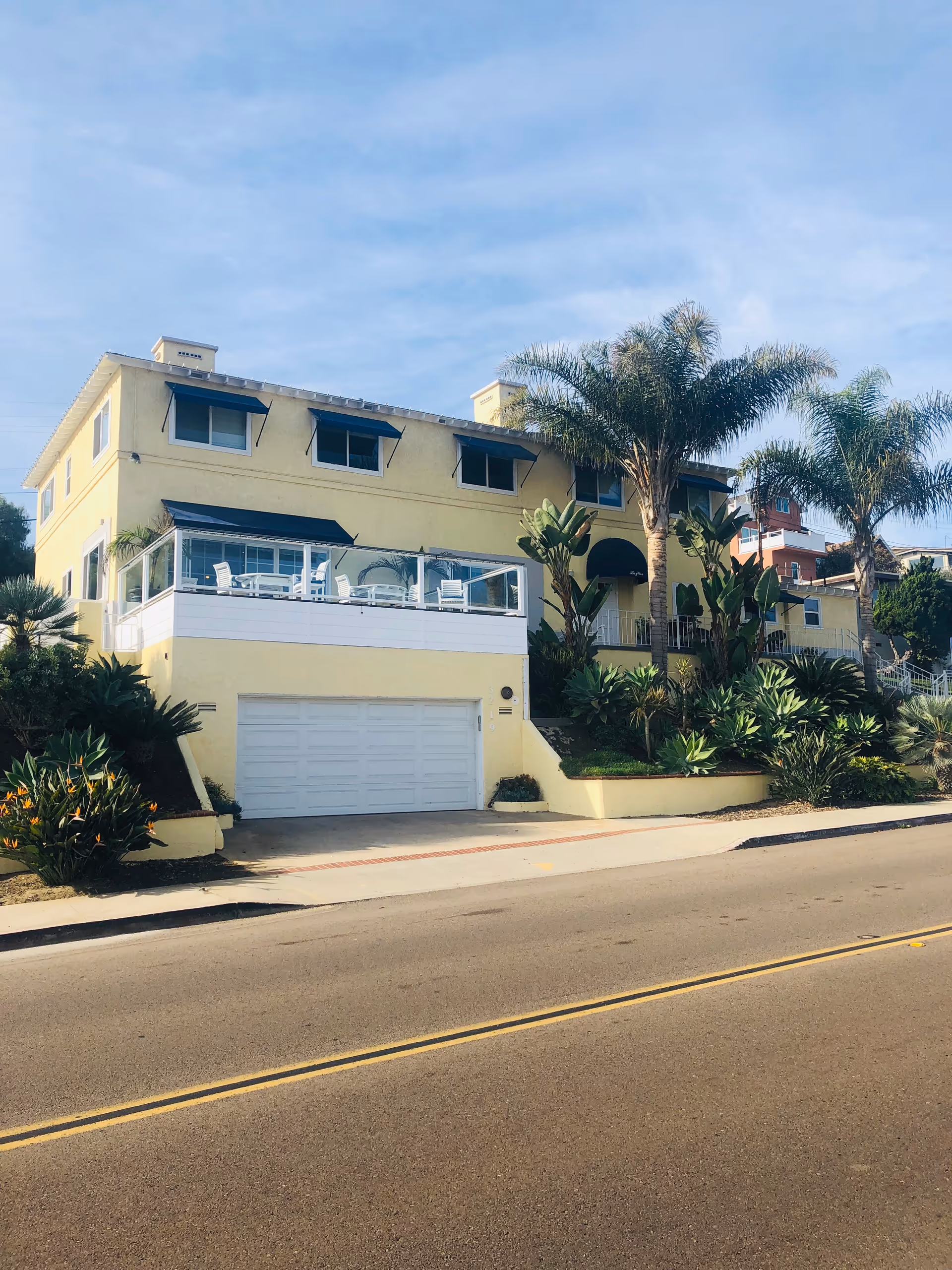 A two-story yellow building with a white garage door and a balcony with white chairs and tables. The building is surrounded by palm trees and other greenery, situated along a paved road under a blue sky.