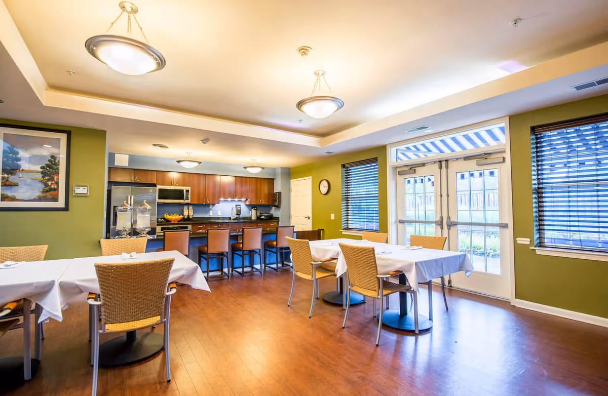 A bright dining area in Arbor Terrace Sudley Manor featuring tables with white tablecloths and wicker chairs. The room has wooden flooring, green walls, and large windows with blinds. In the background, there is a kitchen area with wooden cabinets, stainless steel appliances, and a counter with bar stools. Two ceiling lights illuminate the space.