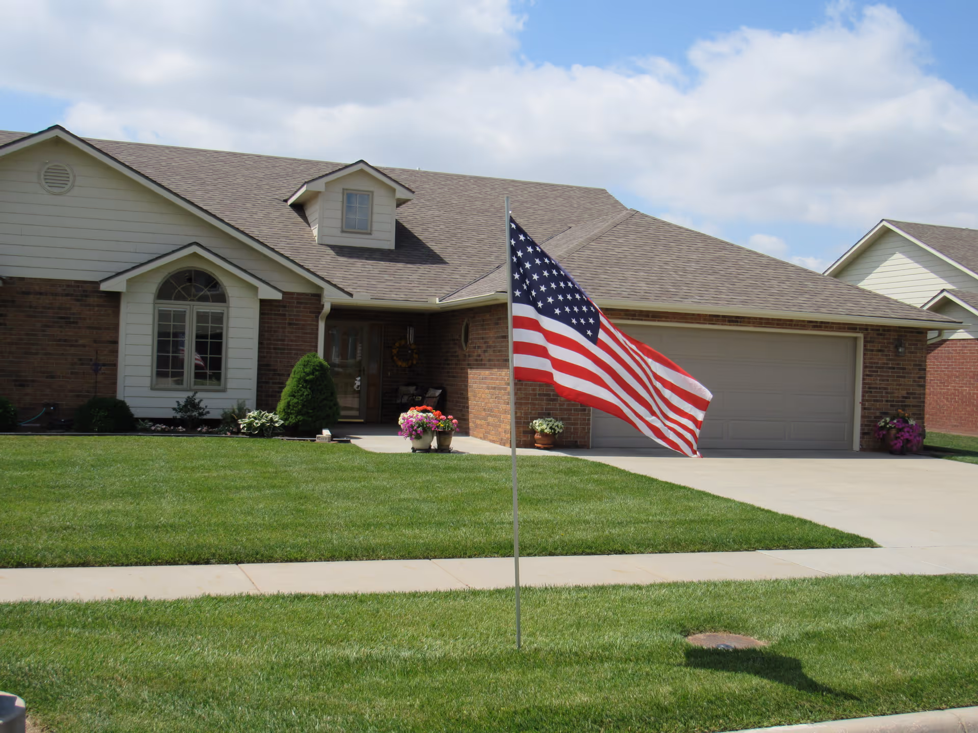 Single-story brick house with a two-car garage, a well-maintained green lawn, and an American flag on a pole in the front yard under a partly cloudy sky.