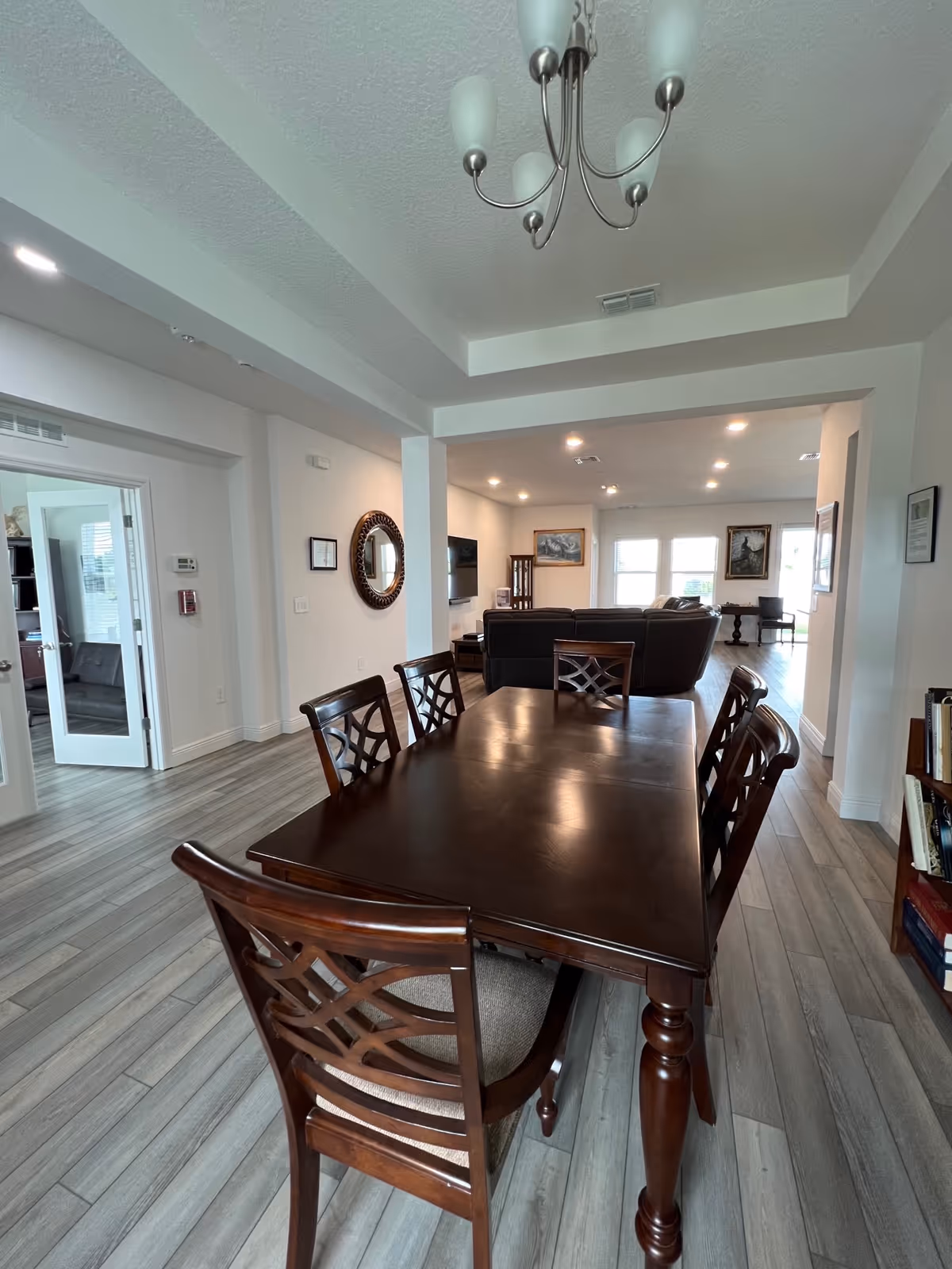 A spacious interior room with a dark wooden dining table and six matching chairs in the foreground. The room has light-colored walls and wood-look flooring. In the background, there is a living area with a dark sofa, a wall-mounted TV, framed artwork, and large windows letting in natural light. A chandelier hangs from the ceiling above the dining table.