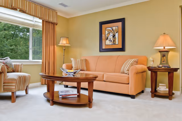 A cozy living room with a peach-colored sofa adorned with striped pillows, a wooden coffee table with a decorative bowl and magazines underneath, a striped armchair near a large window with orange curtains, a floor lamp, and a side table with a lamp and decorative items. A framed abstract painting hangs on the light green wall behind the sofa.