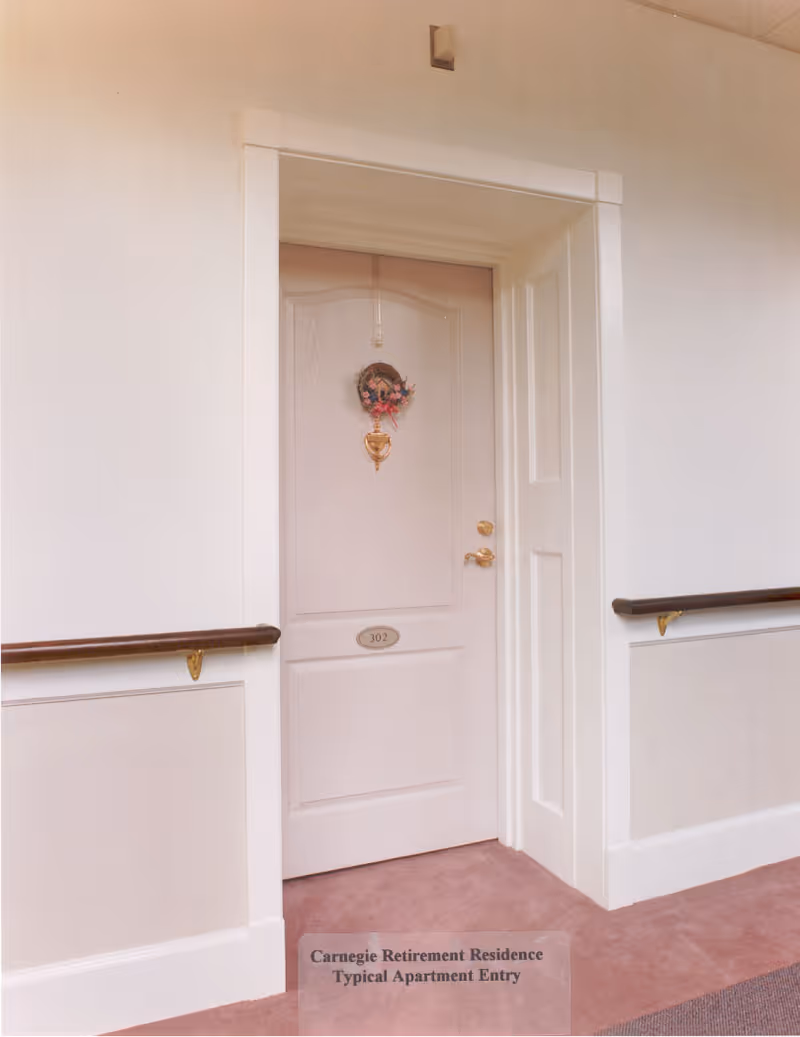 A closed apartment door numbered 302 in a hallway with beige walls and carpeted floor. The door has a decorative wreath and a brass door knocker. Handrails are mounted on both sides of the hallway walls.