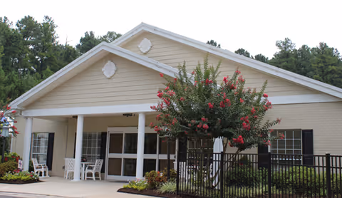 Front entrance of a single-story memory care building with a covered porch, white columns, chairs, and landscaping including a flowering tree.