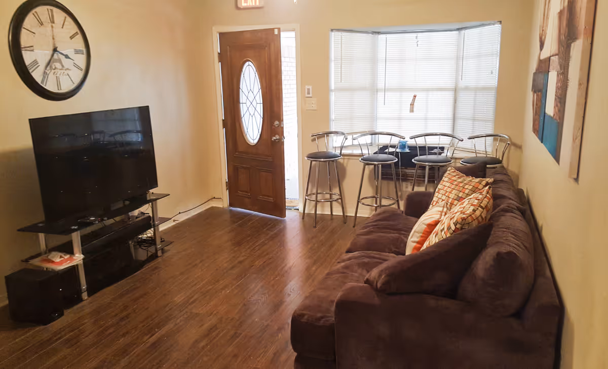 Living room with brown sofa, flat-screen TV, clock, and bar stools by a window.