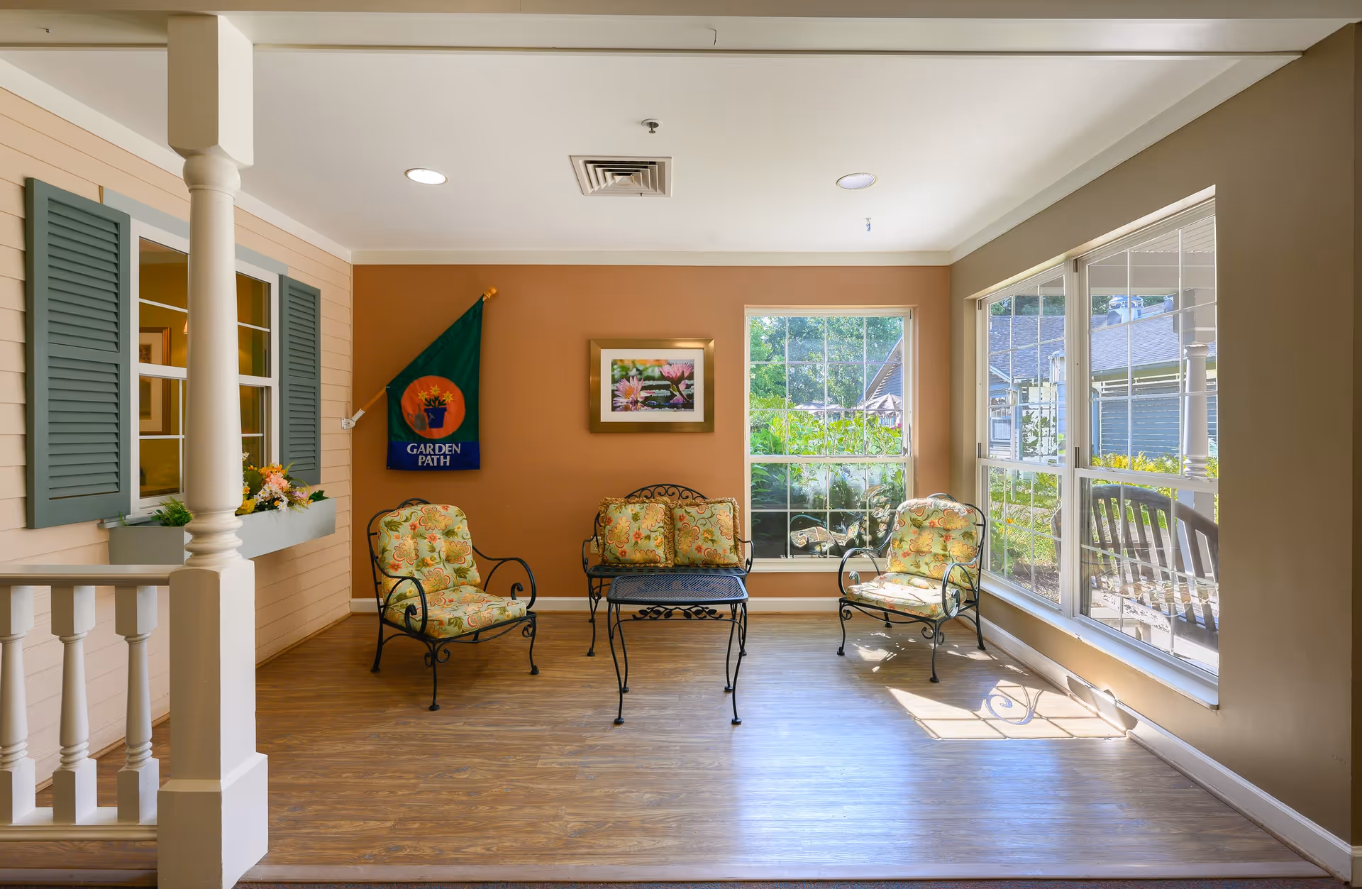 A bright and cozy indoor seating area with three cushioned wrought iron chairs and a matching coffee table. The walls are painted a warm beige color, with a large window letting in natural light and offering a view of greenery outside. There is a decorative flag on the wall that reads 'Garden Path' and a framed picture of flowers. The floor is wood, and there are white railings and window shutters painted in a muted green color.