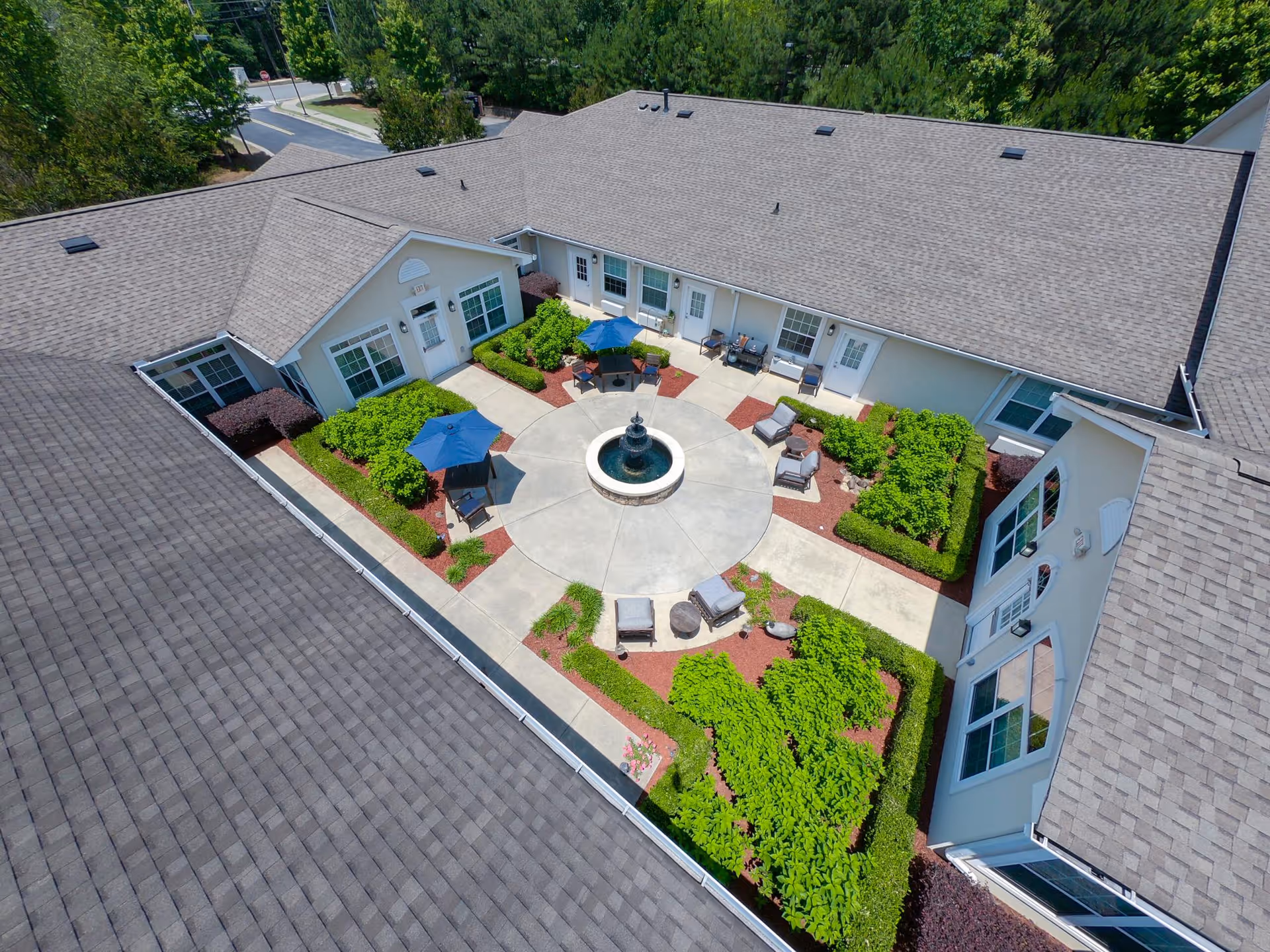 Aerial view of a courtyard within a senior living facility featuring a central circular fountain, surrounded by paved walkways, green bushes, and outdoor seating with umbrellas. The courtyard is enclosed by a building with multiple windows and doors.