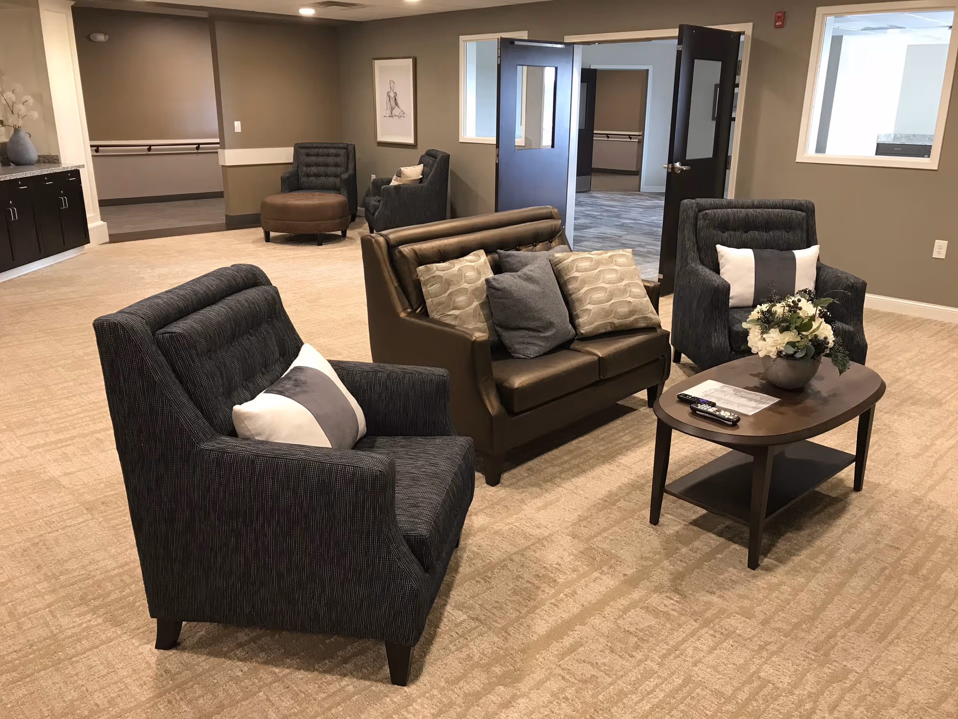 Carpeted common lounge with upholstered armchairs, a leather loveseat, and a coffee table topped with a floral arrangement and remotes.