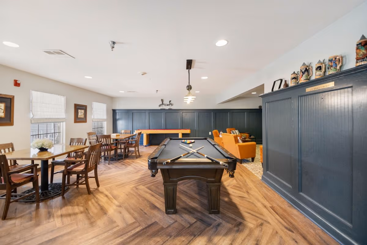 A recreational room with a pool table in the center, two pool cues crossed on the table, and a set of billiard balls. To the left, there are several wooden tables with chairs arranged around them near windows with white blinds. In the background, there is a shuffleboard table and a seating area with orange armchairs and a small table. The room has wood-patterned flooring and dark blue wainscoting on the walls, with decorative steins displayed on a shelf.