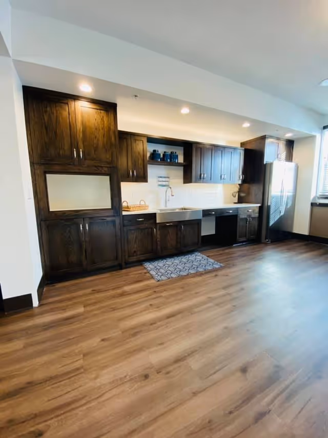 A modern kitchen with dark wooden cabinets, a stainless steel farmhouse sink, a dishwasher, and a stainless steel refrigerator. The kitchen has recessed lighting, a wooden floor, and a small patterned rug in front of the sink. There is a window on the right side letting in natural light.
