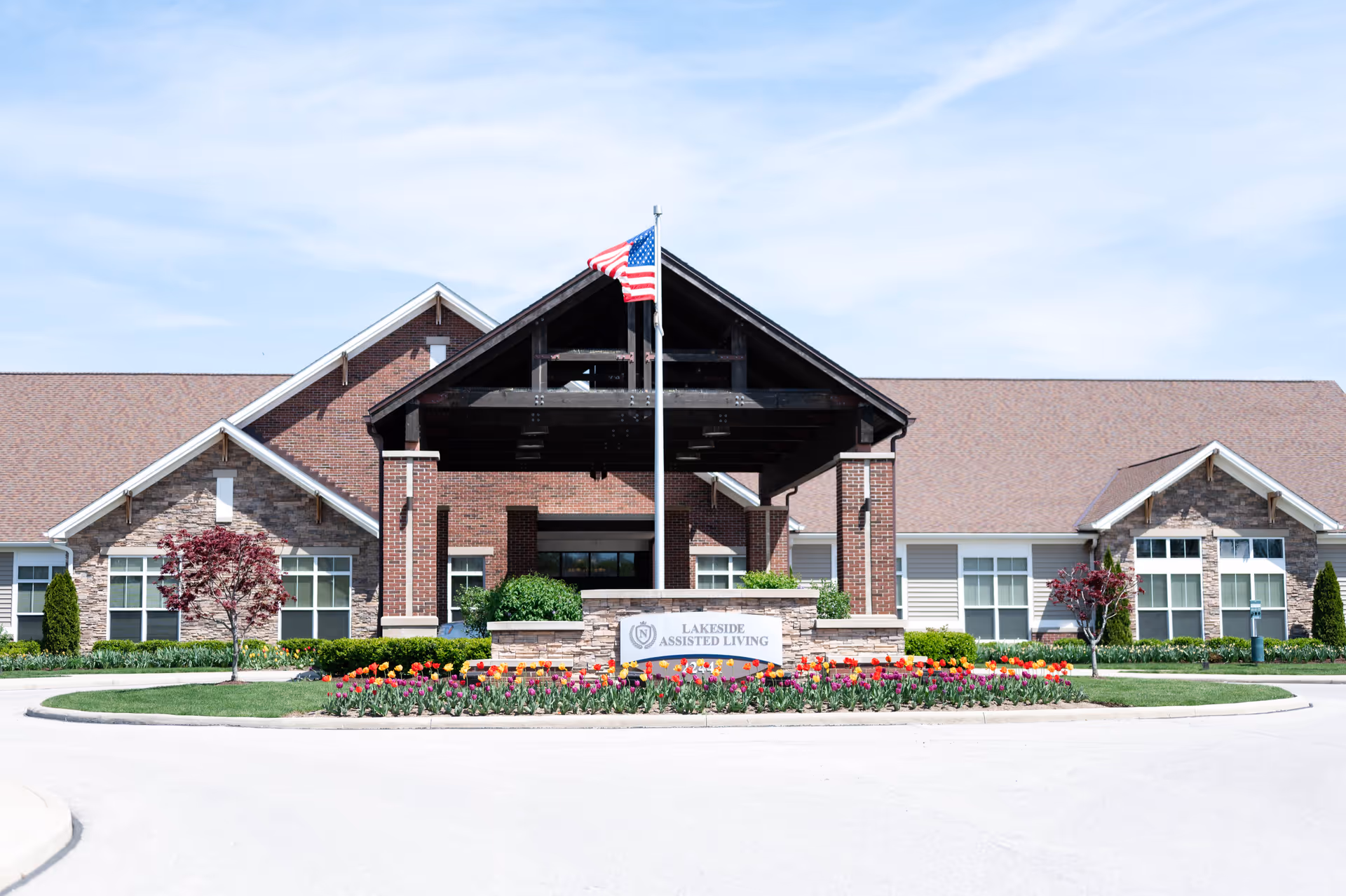 Front entrance of Lakeside Assisted Living building with a covered porte-cochère, an American flag, and colorful flowerbeds.