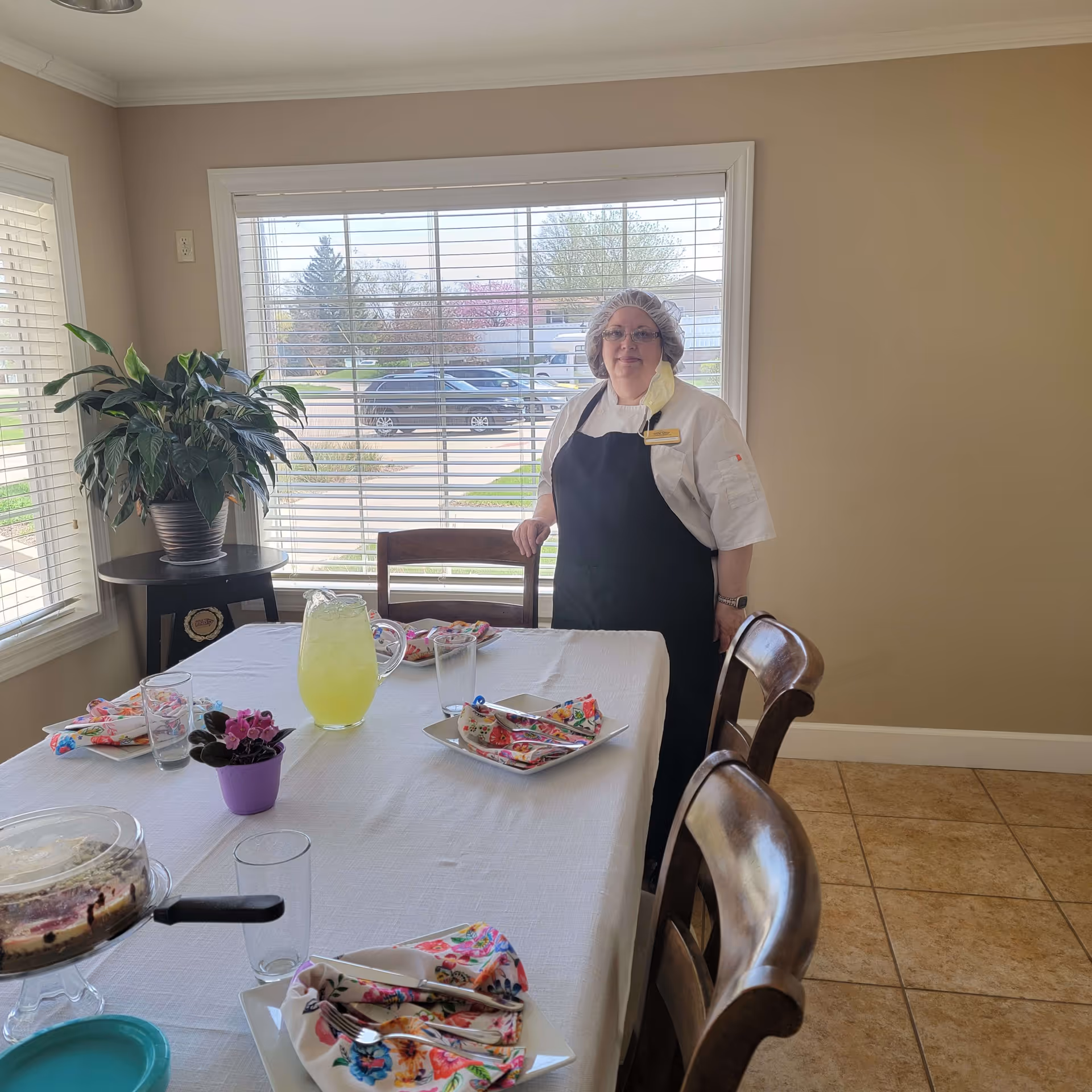 A woman wearing a white shirt, black apron, hairnet, and glasses stands next to a dining table set with colorful napkins, plates, glasses, a pitcher of lemonade, and a cake under a glass cover. The room has beige walls, tiled floor, and large windows with blinds showing a view of parked cars and trees outside. A potted plant is on a small round table near the window.