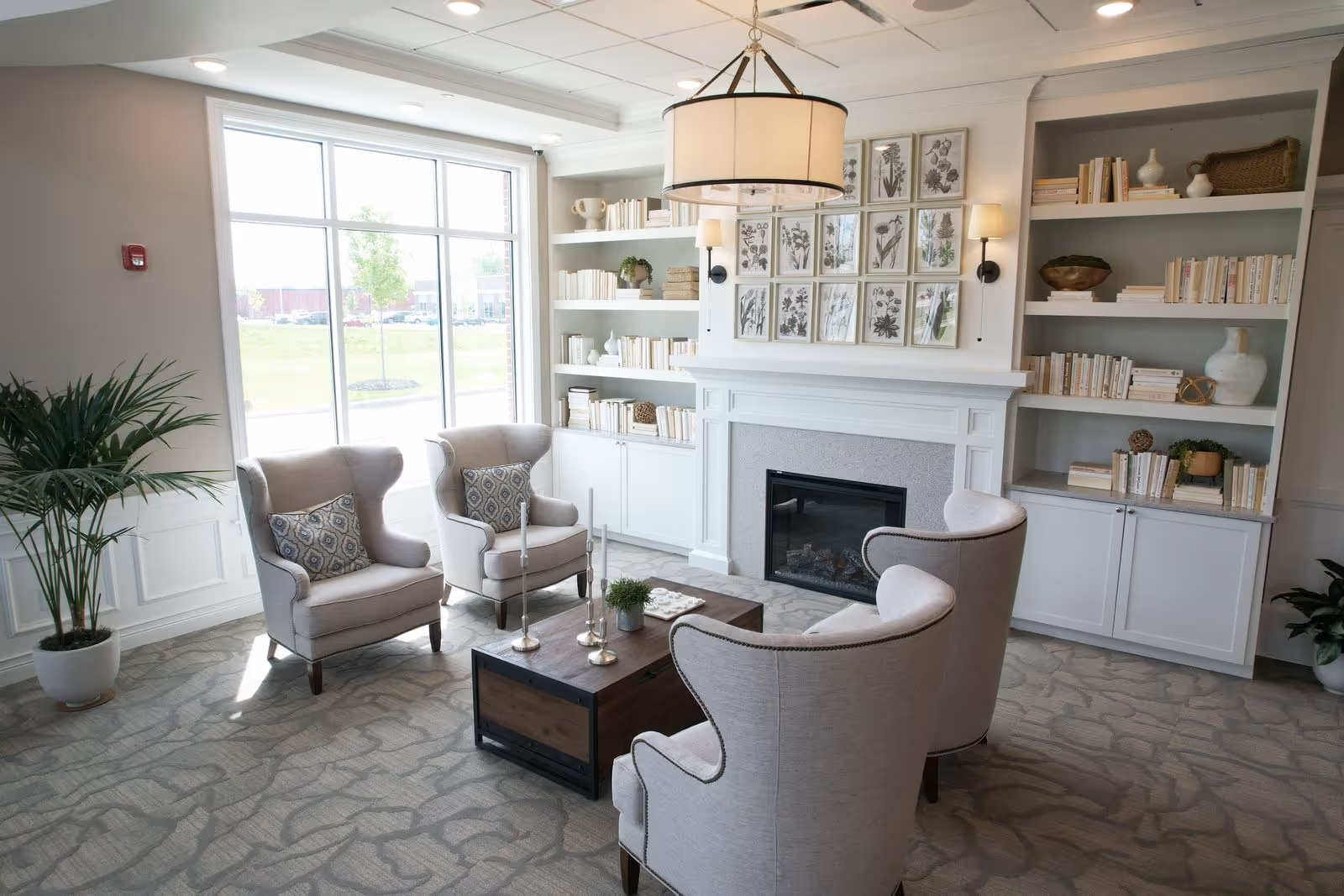 A bright and cozy living room with four beige upholstered armchairs arranged around a wooden coffee table. The room features a large window letting in natural light, built-in white bookshelves filled with books and decorative items, a fireplace with botanical artwork above it, and a large hanging light fixture.
