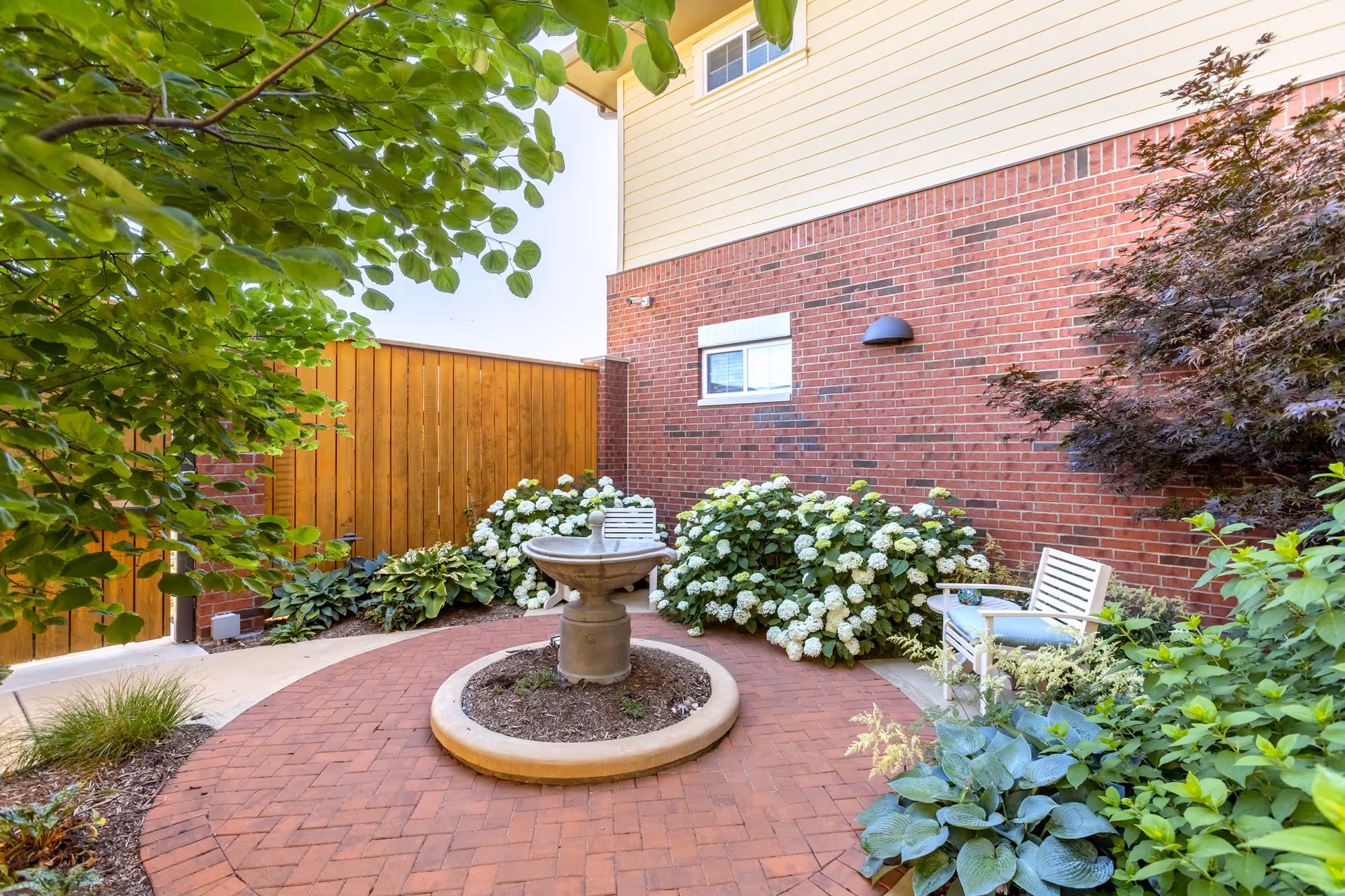 A small outdoor courtyard with a circular brick-paved area featuring a central stone birdbath. The courtyard is surrounded by lush green plants, white flowering bushes, and a wooden fence. There is a white bench with a blue cushion on the right side near a red brick wall of a building.