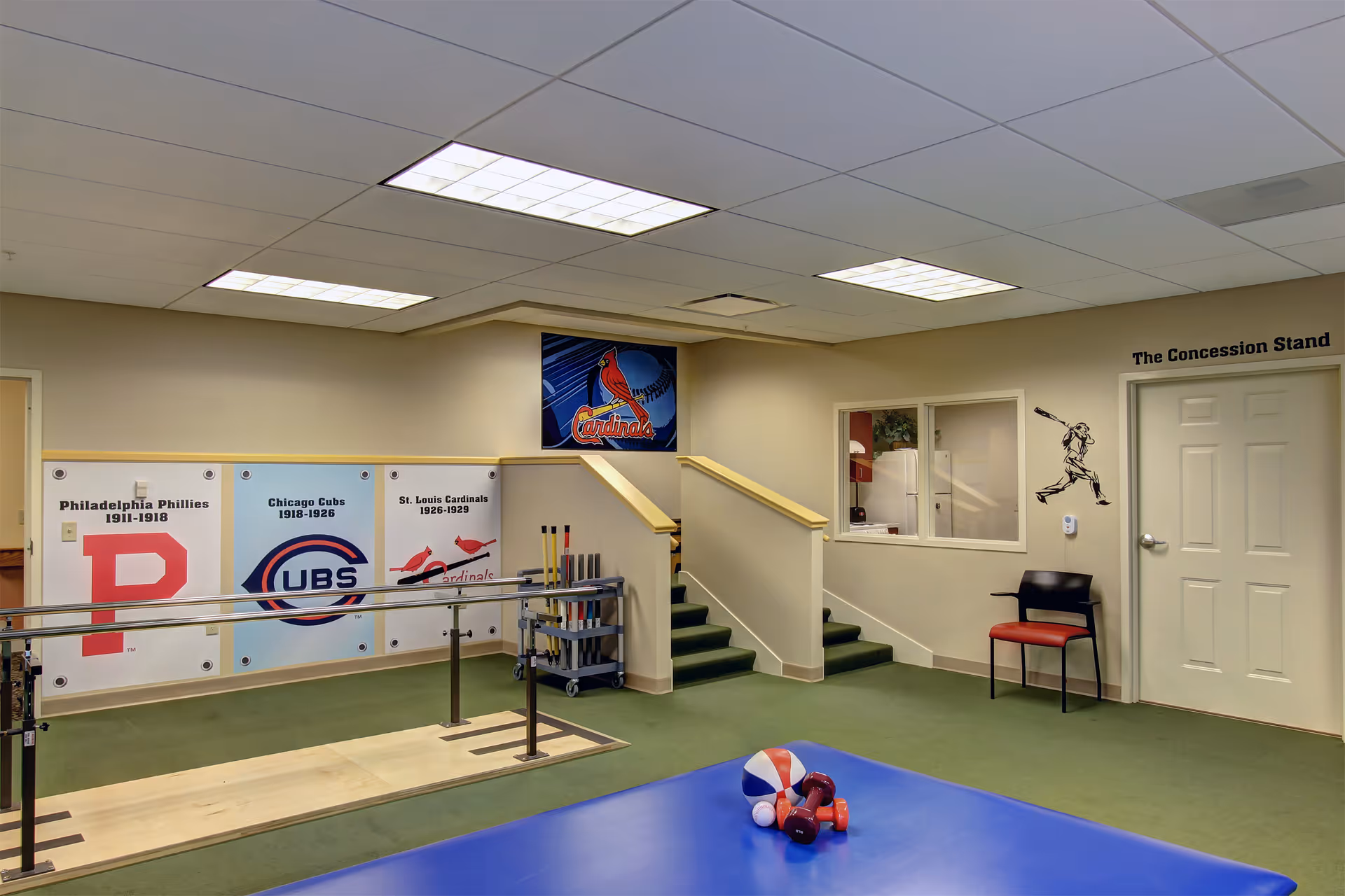 Indoor rehabilitation room with parallel bars for walking exercises, a blue exercise mat with small weights and a ball, a chair, and wall decorations featuring baseball team logos and a baseball player graphic near a door labeled 'The Concession Stand'.