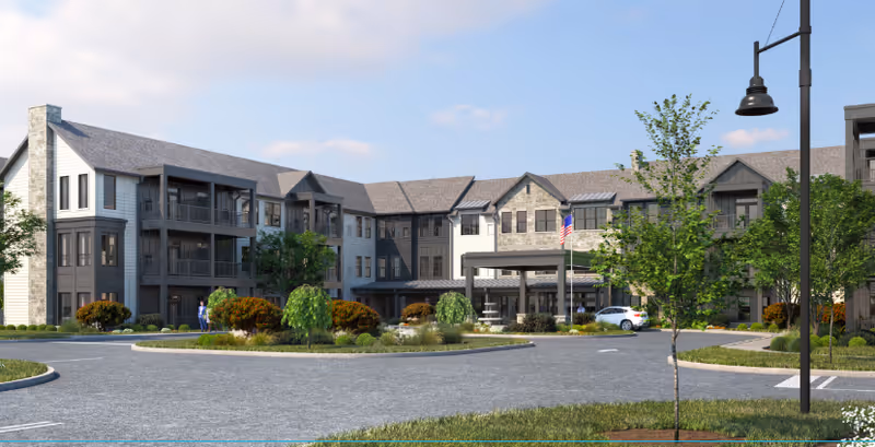 Exterior view of a modern senior living facility building with multiple floors, balconies, and a covered entrance. The building is surrounded by landscaped greenery, trees, and a circular driveway with a parked white car. The sky is partly cloudy.