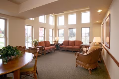 A bright and spacious living room area with large windows letting in natural light. The room features two red sofas, a wicker armchair, a wooden round table with chairs, side tables with lamps, and framed artwork on the wall.