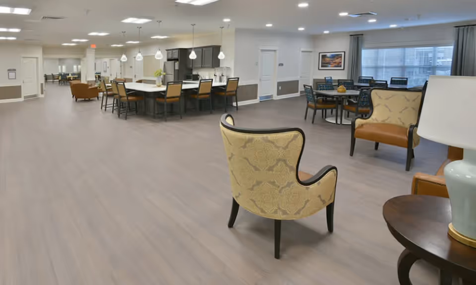 A spacious senior living common area with multiple seating options including patterned armchairs and tables with chairs. The room features a large kitchen island with bar stools, pendant lighting, and a bright window with curtains. The flooring is light wood, and the walls are painted white with wainscoting.