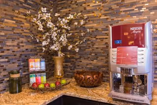 A coffee and tea station with a Nescafe Alegria coffee machine, a variety of tea bags in a display box, a decorative vase with white flowers, a wooden bowl, and some apples on a granite countertop against a textured stone tile wall.