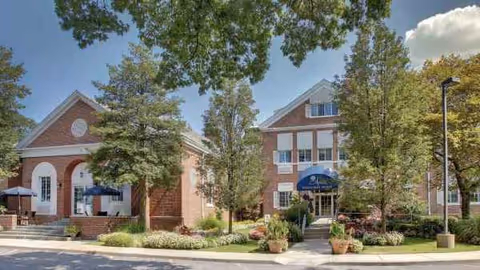 Brick senior living facility exterior with trees, planters, and an entrance under a blue awning.