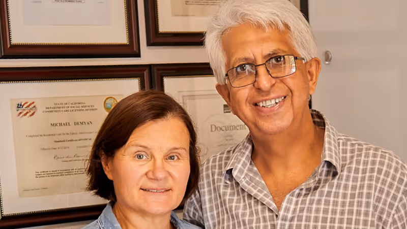 A smiling middle-aged man with gray hair and glasses stands next to a middle-aged woman with short brown hair. Behind them on the wall are several framed certificates and documents.